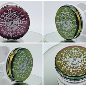 May include: A white phone grip with a round, iridescent, green and gold skull design. The skull is surrounded by a white spiderweb design.