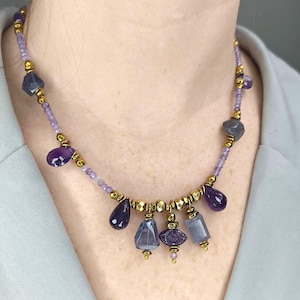 May include: A necklace featuring a series of purple amethyst stones in various shapes, including teardrops and faceted beads, interspersed with small gold-toned metallic beads and light purple seed beads, arranged in a multi-strand style design.