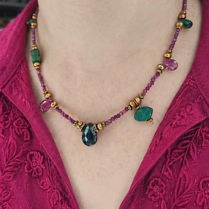 Puede incluir: Un collar que presenta una hilera de pequeñas cuentas moradas intercaladas con espaciadores metálicos dorados. La pieza está decorada con varios dijes colgantes más grandes en forma de gema en tonos azul profundo, verde y rosa.