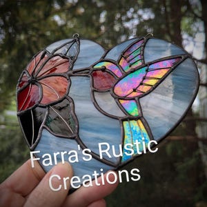 May include: Heart-shaped stained glass art featuring a hummingbird and flower. The hummingbird has iridescent wings, a red throat, and a long beak. The flower is red and orange. The text "Farra's Rustic Creations" is visible.