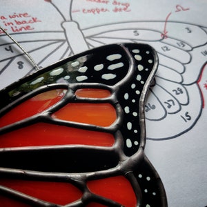 Könnte beinhalten: Ein Buntglas-Schmetterling mit schwarzen, roten und weißen Akzenten. Der Schmetterling ist teilweise zusammengebaut und hat einen Draht daran befestigt. Das Bild zeigt auch ein Diagramm eines Schmetterlings mit nummerierten Abschnitten.