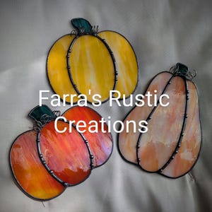 May include: Three stained glass pumpkin decorations in orange, yellow, and peach. Each pumpkin has a dark metal outline and a green stem. The text "Farra's Rustic Creations" is displayed in white.