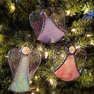 May include: Three stained glass angel ornaments hanging on a Christmas tree. The angels are made of clear glass with iridescent accents and have silver foil wings. The angels are in shades of pink, purple, and green.