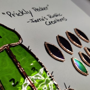 Op de afbeelding: Een glas-in-lood cactusontwerp met groene en koperen accenten. De afbeelding bevat de tekst "Prickly Pecker" en "-Jarra's Rustic Creations". Er zijn ook verschillende bladvormige glazen stukken aanwezig.