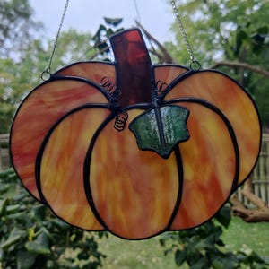 May include: A stained glass pumpkin with a green leaf and orange, yellow, and red glass. The pumpkin is hanging from a silver chain.