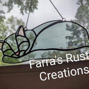 May include: A stained glass cat silhouette in a sleeping pose, outlined in black. The cat's ears and nose are accented with pink and beige. The artwork is suspended by a chain and hangs in front of a window. The text "Farra's Rustic Creations" is visible.