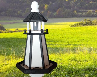 May include: A black and white wooden lighthouse bird feeder with a white post. The feeder has a black roof and a white base. The feeder is in a field of green grass.
