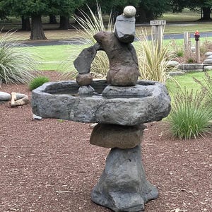 May include: A stacked stone fountain with a basin and a unique, artistic design. The fountain features a series of irregularly shaped stones, creating a natural and textured appearance. The water feature is gray and brown, and is set in a garden setting.