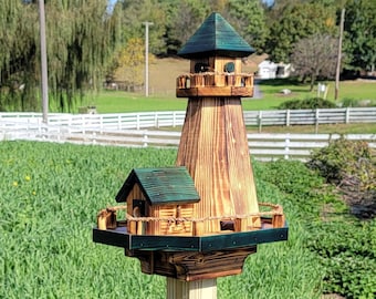 May include: A wooden birdhouse shaped like a lighthouse with a green roof and a brown base. The birdhouse is mounted on a wooden pole and is surrounded by green grass.
