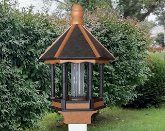 May include: A wooden bird feeder with a pagoda-style roof. The roof is black and brown, and the feeder is supported by a white post. The feeder is designed to attract birds to a garden or outdoor space.