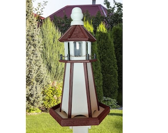 May include: A white and brown wooden lighthouse bird feeder with a black metal railing and a white post. The feeder is in a grassy yard with green trees in the background.