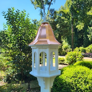May include: A white bird feeder with a copper roof and a decorative finial. The feeder has a hexagonal base and a tall, slender post. The feeder is surrounded by green foliage.