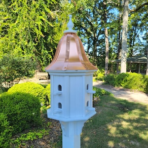 May include: A white birdhouse with a copper roof and a decorative finial. The birdhouse has multiple nesting compartments and is mounted on a white post.