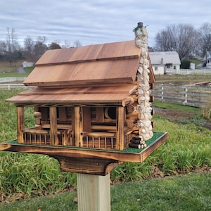 May include: A wooden birdhouse shaped like a log cabin with a stone chimney. The birdhouse has a green roof and a small porch. It is mounted on a wooden post.