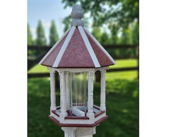 May include: A red and white bird feeder with a decorative roof and a clear plastic feeding area. The feeder is mounted on a white pole.