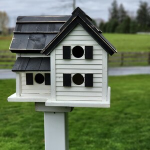 May include: A white birdhouse with a black roof and black accents. The birdhouse has two nesting compartments with round openings and is mounted on a white pole.