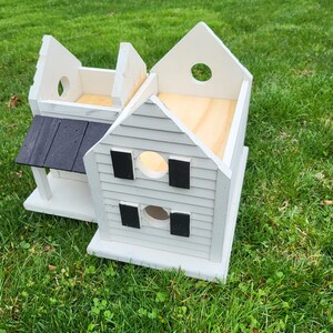 May include: A white wooden birdhouse with a black roof and black accents. The birdhouse has a small porch and multiple round openings.