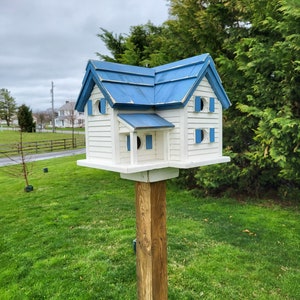 May include: A white birdhouse with a blue roof, featuring multiple windows and a small porch. The birdhouse is mounted on a wooden post in a grassy yard.