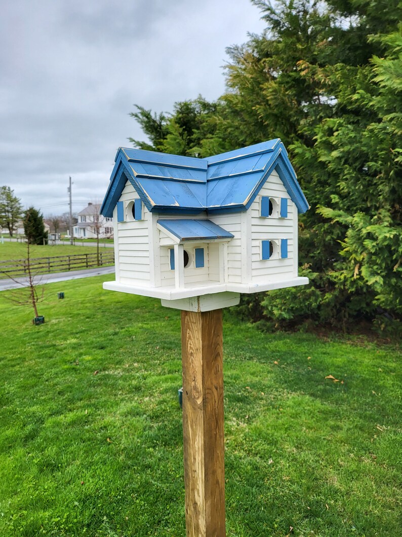 May include: A white birdhouse with a blue roof, featuring multiple windows and a small porch. The birdhouse is mounted on a wooden post in a grassy yard.
