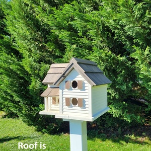 May include: A white birdhouse with a brown stained roof, two entry holes, and a white post. The birdhouse is in front of a green bush.