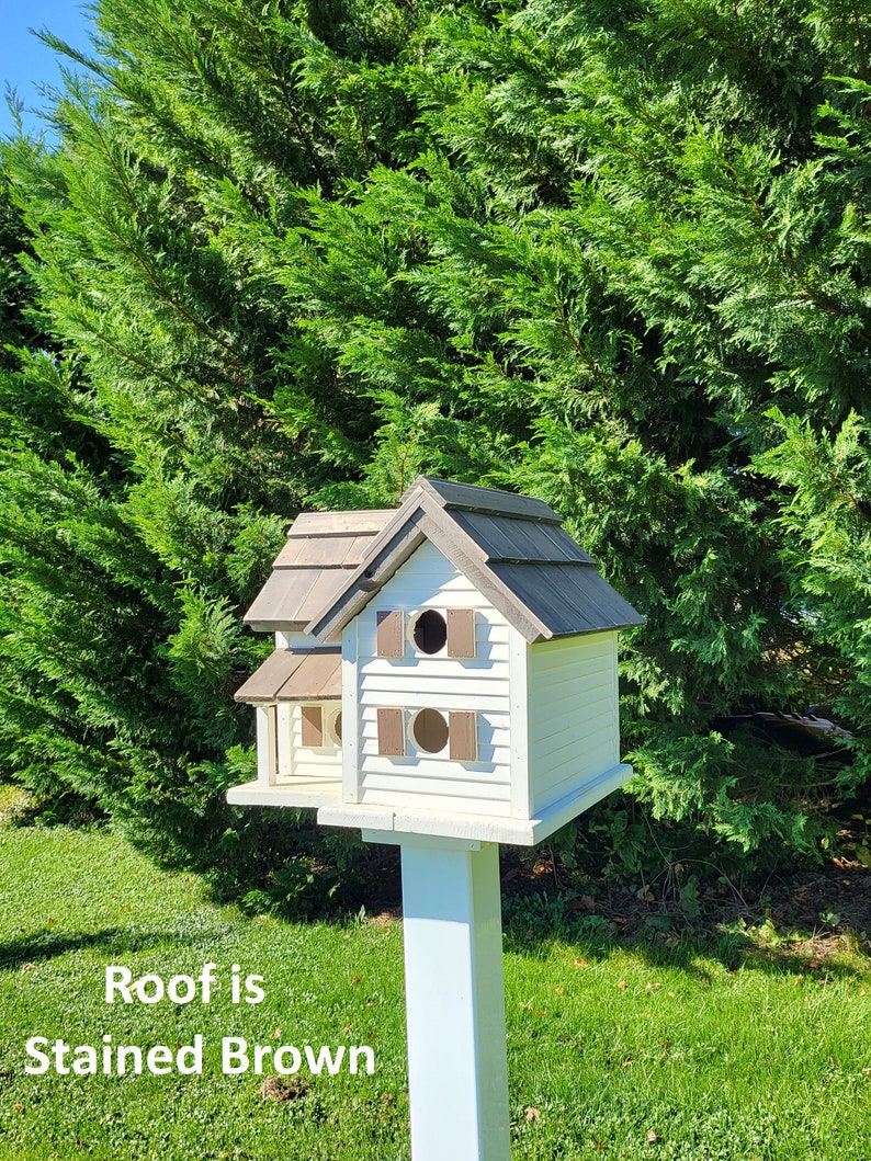 May include: A white birdhouse with a brown stained roof, two entry holes, and a white post. The birdhouse is in front of a green bush.