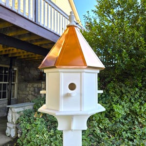 May include: A white birdhouse with a copper roof, mounted on a white post. The birdhouse has a small, round entrance hole and a decorative finial on top.
