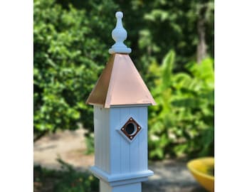 Puede incluir: Casa de pájaros blanca con techo de cobre y un agujero de entrada en forma de diamante. La casa de pájaros está montada en un poste blanco.