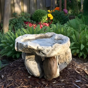 May include: A gray stone bird bath with a shallow basin and a rough, textured surface. The bird bath is supported by three large stones and sits in a garden setting with green plants and colorful flowers.
