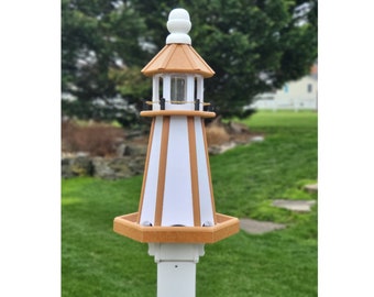 May include: A white and tan lighthouse-shaped bird feeder with a white base and a tan roof. The bird feeder has a small window and is decorated with black chains. It is set on a white pole and surrounded by green grass.
