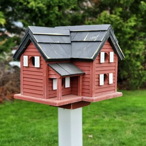 May include: A red wooden birdhouse with a black roof and white accents. The birdhouse is mounted on a white pole and is surrounded by green grass.