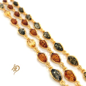 Op de afbeelding: Een gouden kettingarmband met drie rijen ovale amber kralen. De kralen zijn een mix van bruin, geel en groen amber.