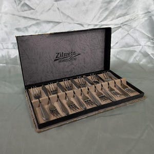 Può includere: Un set di forchette argentate in una scatola nera con il marchio "Zilmeta" stampato sul coperchio. Le forchette sono disposte ordinatamente in slot individuali all'interno della scatola, suggerendo un set da pranzo formale.