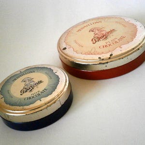 Może przedstawiać: Dwa okrągłe metalowe puszki z pokrywkami. Puszka po lewej stronie ma niebieską pokrywkę i napis "Baronie Mleczna Czekolada" na etykiecie. Puszka po prawej stronie ma czerwoną pokrywkę i napis "Baronie Czekolada Gorzka" na etykiecie.