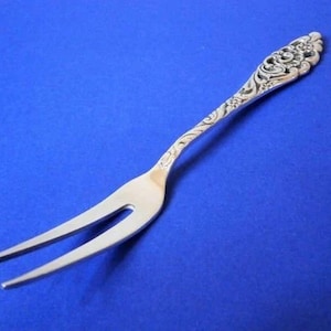 Può includere: Una forchetta da portata argentata con un design floreale ornato sul manico. La forchetta ha due rebbi ed è lunga circa 18 cm.