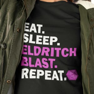 Könnte beinhalten: Schwarzes T-Shirt mit weißem Text, der "Essen. Schlafen. Eldritch Blast. Wiederholen." lautet und einem lila und schwarzen Dungeons and Dragons Würfel-Symbol.
