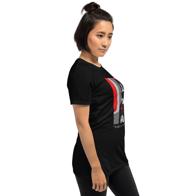 May include: A black short-sleeved t-shirt featuring a graphic print on the side. The design displays a stylized figure in black, white, and grey tones, accented by a prominent red cross and vertical red stripes, with Japanese text at the bottom.