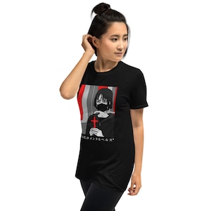 May include: A black short-sleeve t-shirt featuring a graphic print of a person with short dark hair wearing a black face mask and holding a red cross. The design includes red and grey vertical stripes and Japanese text at the bottom.