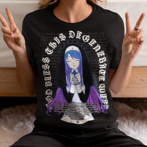 May include: Black t-shirt with a graphic of a nun with purple and blue accents. The text on the shirt reads "God bless this legend embrace wtf".