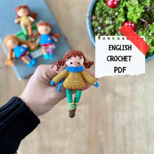 May include: Crocheted doll with brown hair, a yellow sweater, a blue scarf, and green trousers. The doll is holding a small blue scarf. The image also includes a piece of paper with the text "ENGLISH CROCHET PDF".