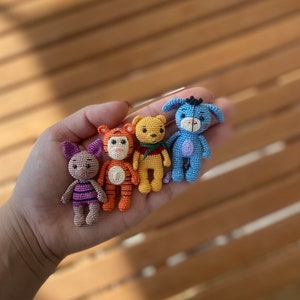 May include: A hand holding four small crocheted stuffed animals. The animals are Winnie the Pooh, Tigger, Eeyore, and Piglet. The animals are all different colors and have different facial expressions.