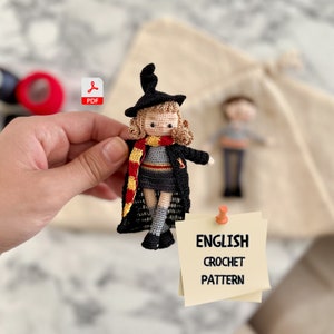 May include: Crochet pattern for a small doll wearing a black witch's hat, a grey and red scarf, and a black dress. The doll has blonde hair and brown eyes. The pattern is available as a PDF download. The text on the note reads "ENGLISH CROCHET PATTERN".