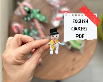 Może przedstawiać: Pluszowa zabawka amigurumi w kształcie bałwana z czarnym kapeluszem, żółtym szalikiem i niebieskimi rękawiczkami. Zabawka jest trzymana przez rękę. Na tle widoczna jest notatka z tekstem "ENGLISH CROCHET PDF".
