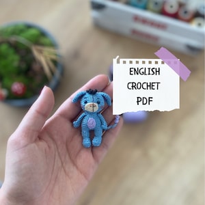 Könnte beinhalten: Ein kleiner blauer gehäkelter Esel mit violetten Akzenten. Der Esel wird in einer Hand gehalten. Im Hintergrund befindet sich ein Zettel mit dem Text "ENGLISH CROCHET PDF".
