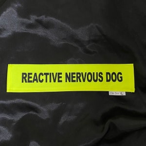 May include: A neon yellow patch with the text "REACTIVE NERVOUS DOG" in black. The patch is rectangular and has a small logo in the bottom right corner. The background is a black, shiny fabric.