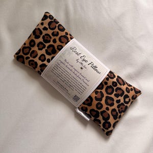 May include: A rectangular eye pillow with a leopard print design. The pillow has a brown and black spotted pattern and a label that reads "Rest Eye Pillow by ellie bee". It is made with organic lavender buds from Provence.