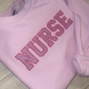 Puede incluir: Sudadera rosa con la palabra "NURSE" bordada en letras de purpurina rosa.