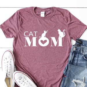 May include: A maroon t-shirt with the text "CAT MOM" in white. The "O" in "MOM" is a heart shape. Two white cat silhouettes are above the text. White sneakers and blue jeans are next to the shirt.