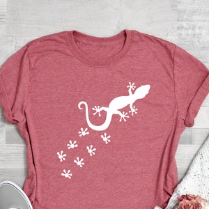 May include: A maroon t-shirt with a white gecko design and a trail of footprints. The gecko is facing right and has a long tail.