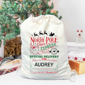 May include: A white canvas Santa sack with a red, green, and black North Pole Express design. The sack has a drawstring closure and the text "Special Delivery for Audrey" is printed on the front. The text "Do Not Open Until December 25th" is printed below the name.
