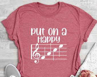May include: A red t-shirt with the text "Put on a Happy" and a treble clef and musical notes on a staff printed in white.